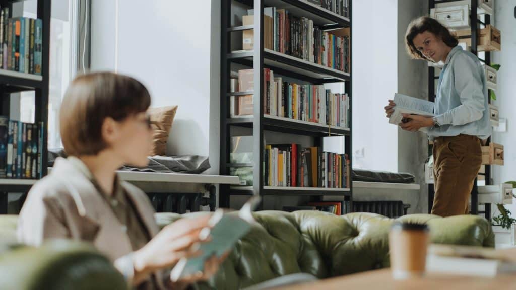 A man stands by a tall bookshelf reading, while a blurred person sits on a green sofa.