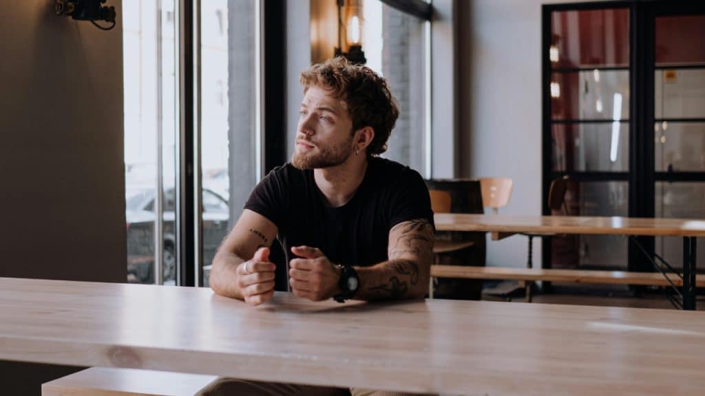 A man with tattoos and a nose ring sits alone at a long wooden table.