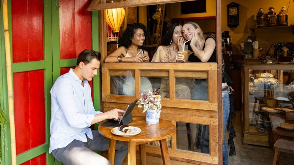 A man uses a tablet at a table while three women talk in a window behind him.