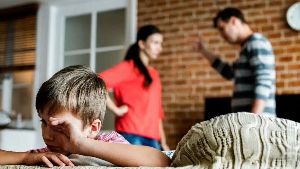 A young boy rubs his eyes in the foreground while two adults argue in the background.