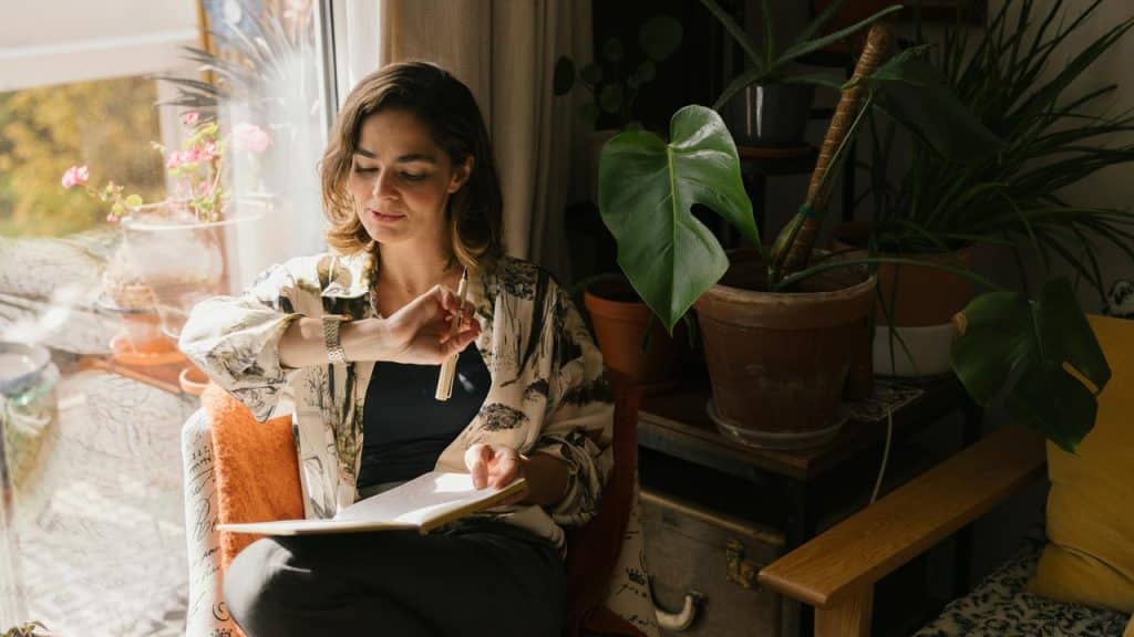 A woman sits by a window, checking her watch while holding a notebook.