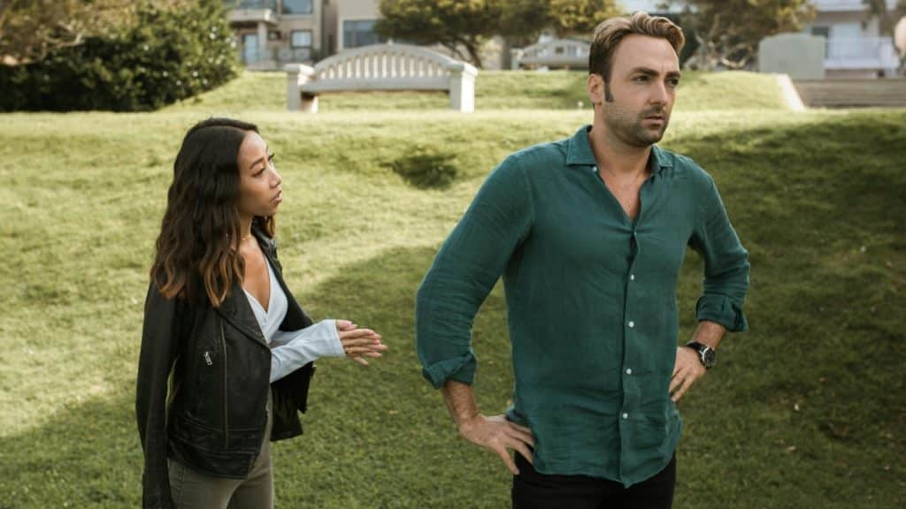 A woman in a leather jacket reaches toward a man who is walking away on a grassy hill.