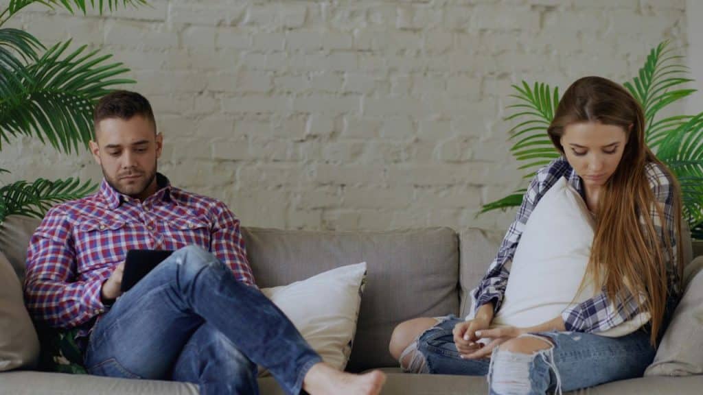 A man and woman sit on a couch, looking away from each other in silence.