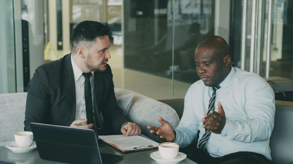 Two men in business attire sit at a table with coffee and a laptop, talking.