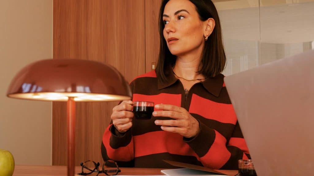 A woman holding a cup of coffee while looking to the side at a desk with a laptop.