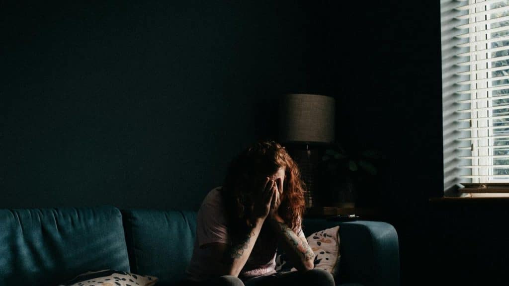 A person sitting on a couch with their hands covering their face in a dimly lit room.