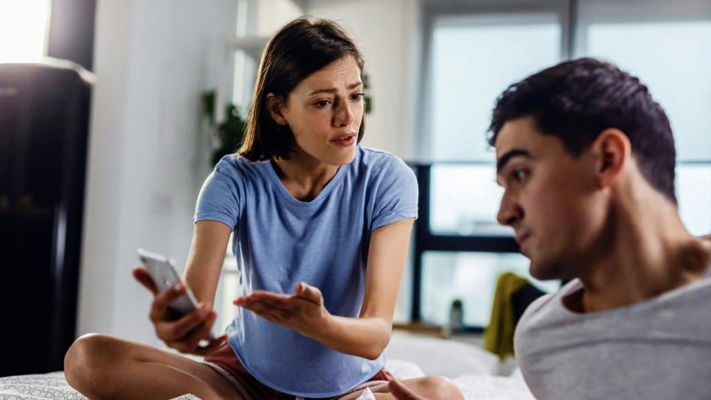 A woman holding a smartphone talks to a man while they both sit on a bed together.