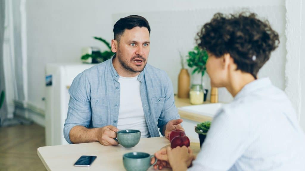 A man sitting at a table looks toward a person and speaks with a tense expression.