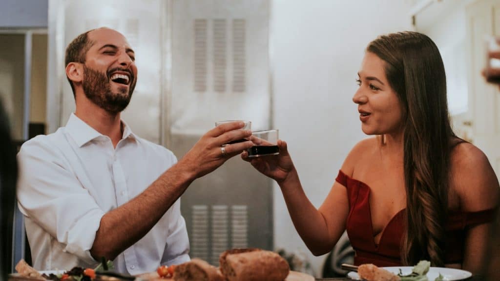 A man laughing and a woman smiling while clinking small glasses over a dinner table.