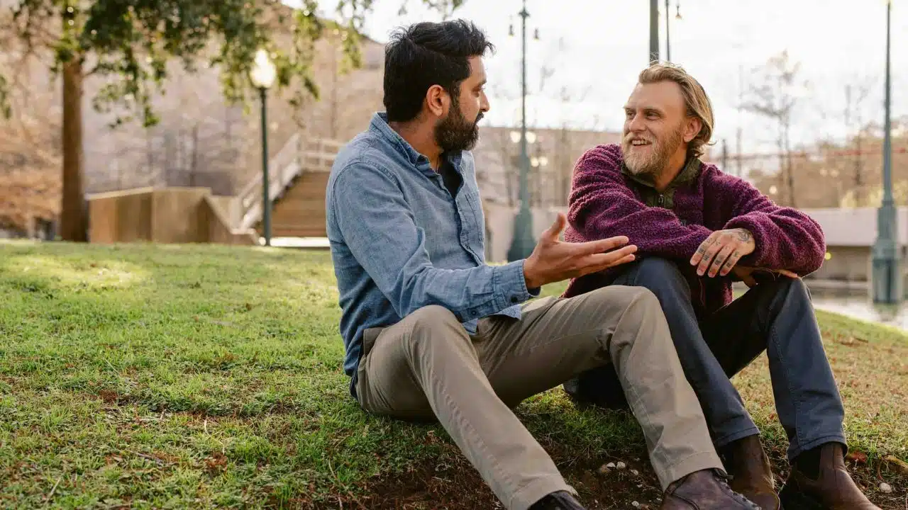 Two men sit on a grassy hill having a conversation while one gestures with his hand.