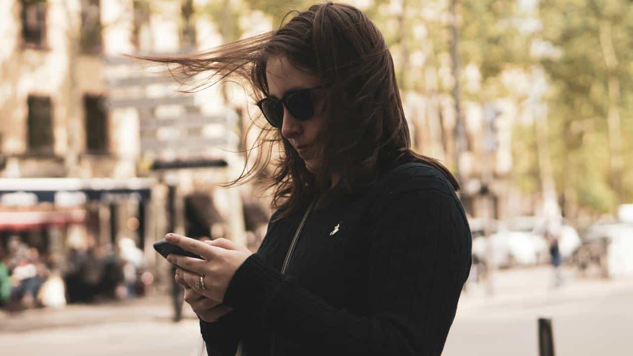 A woman wearing sunglasses checking her phone on a city street.