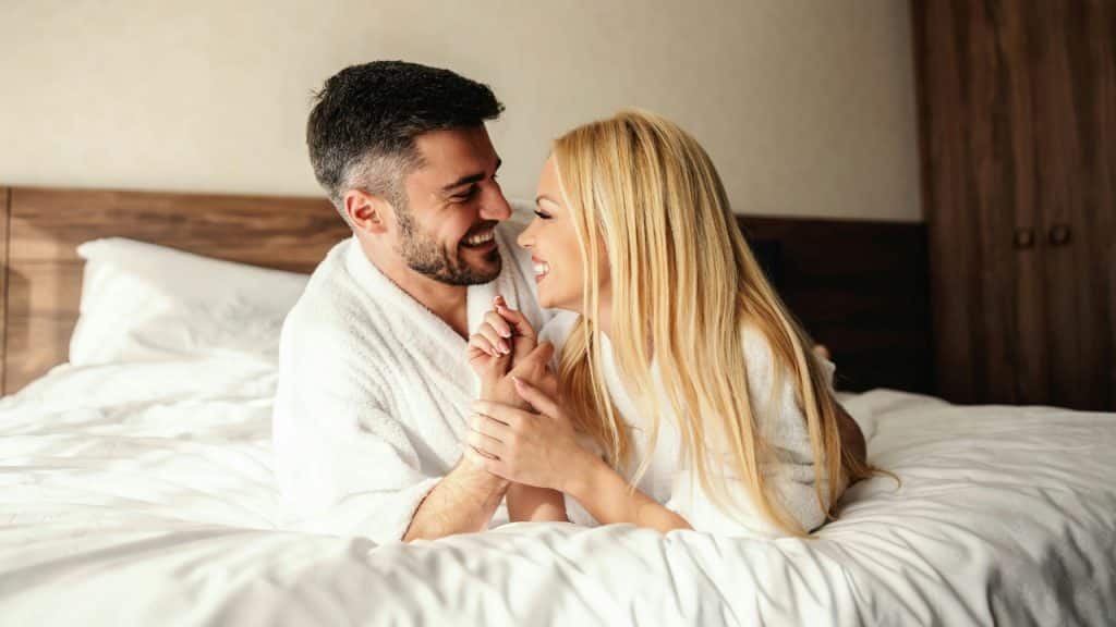 A man and woman in white robes smile at each other while lying in bed.