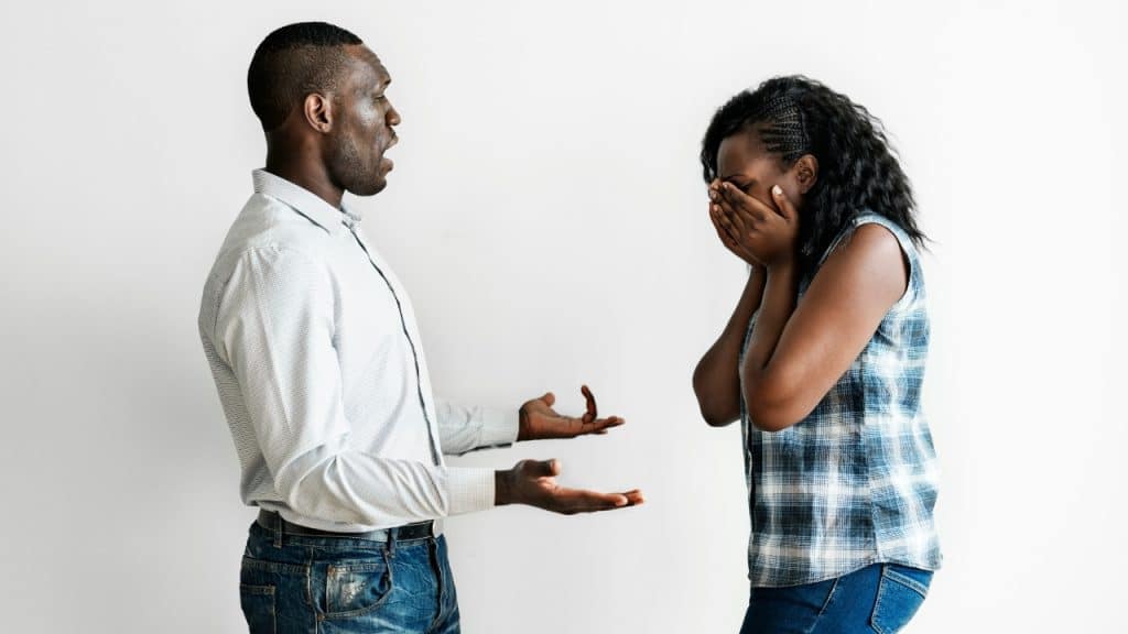 A man gestures with open hands while a woman stands nearby covering her face with hands.