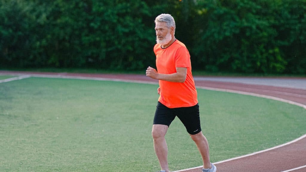 A man with a white beard runs on a green athletic track wearing an orange shirt.