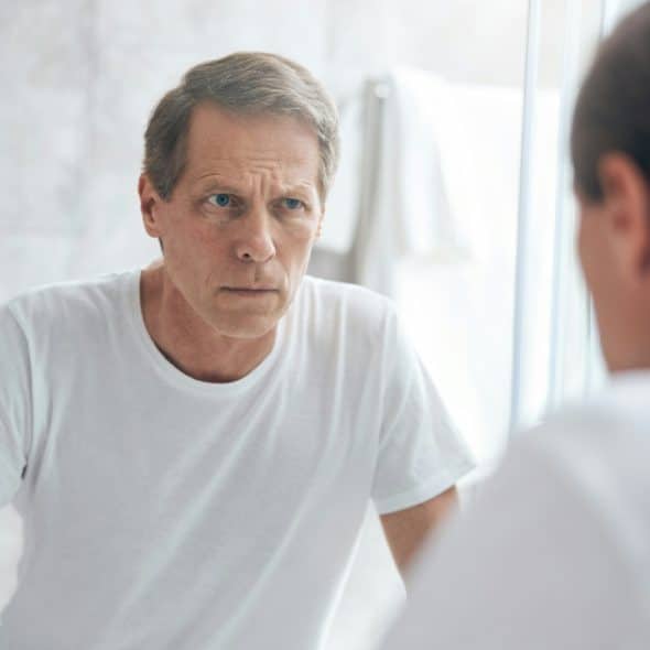 A man in a white t-shirt looks at his reflection in a bathroom vanity mirror.