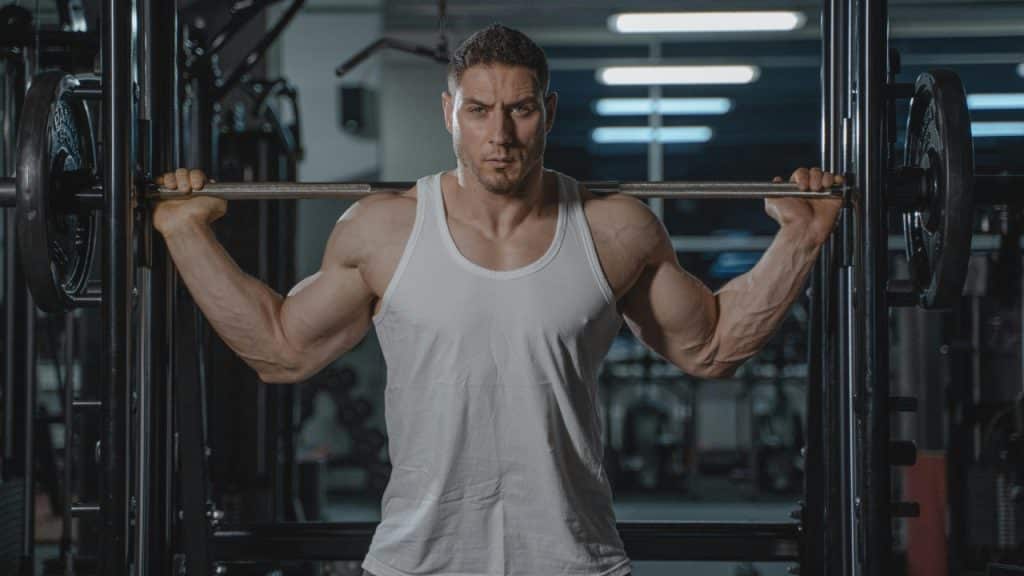 A muscular man in a white tank top looks forward while holding a barbell at a gym.