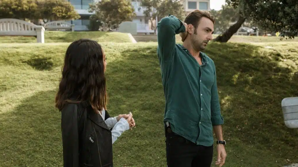A man and woman stand on a grassy hill, and the man has his hand on the back of his neck, looking away.
