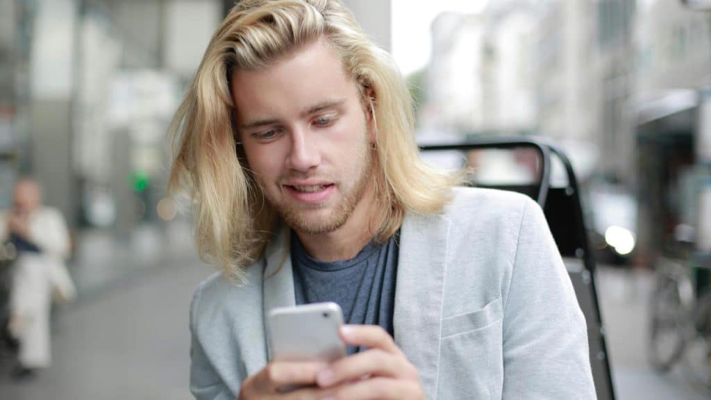 A long-haired man in a light blazer looking at his smartphone while sitting outdoors in a city.