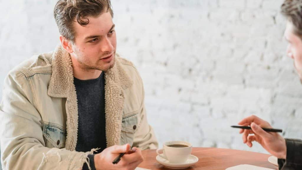 A man in a shearling jacket discussing papers over coffee at a table.