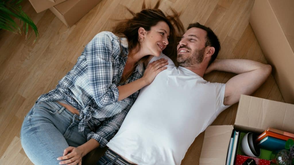 A couple lies on a wooden floor together near several cardboard boxes.