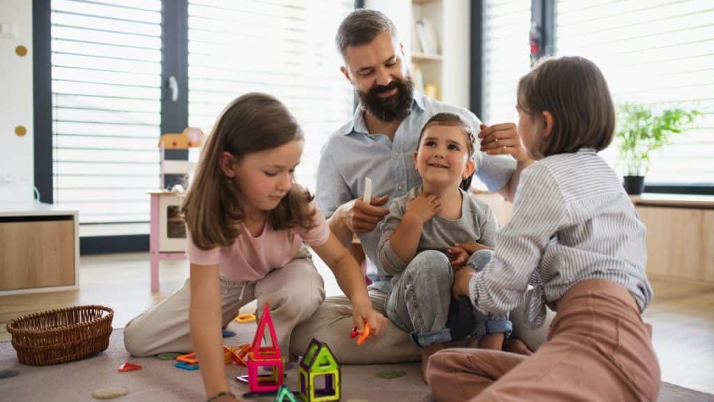 A bearded man sits on a rug playing with three young children and toys.