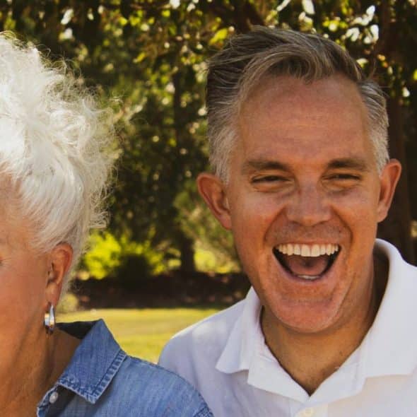 An older couple laughing together under the shade of trees.