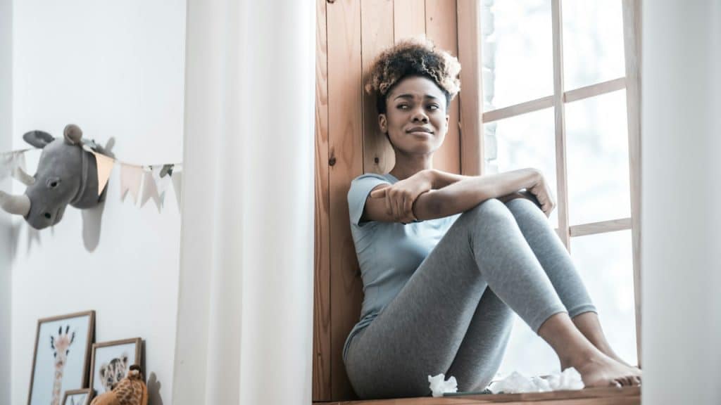 A depressed young dark-skinned woman, barefoot in home clothes, sitting on a windowsill looking into the room.