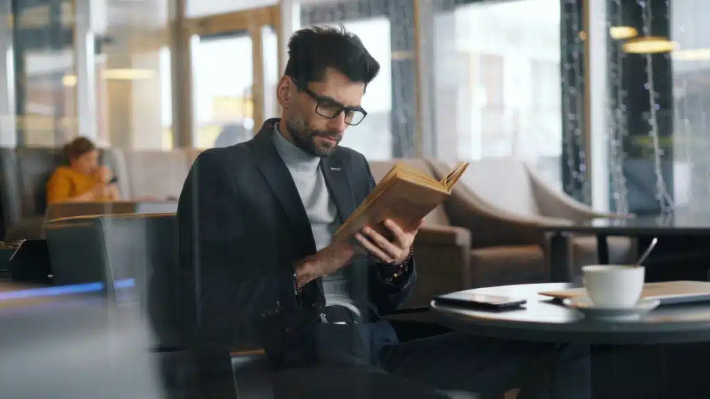 A man with glasses and a blazer sits in a cafe while reading a hardcover book.
