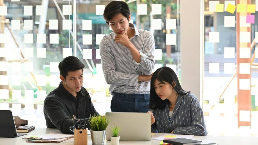 Three people sit and stand around a laptop while working in an office.