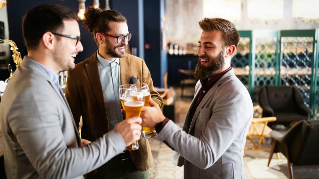 Three men in business casual attire smile and toast with glasses of beer.