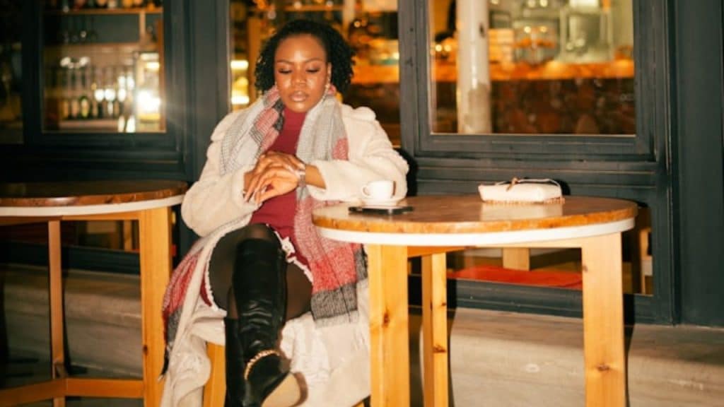 A woman wearing a scarf sits at a cafe table checking her wristwatch.