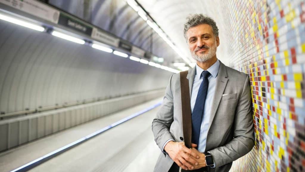 A smiling man in a grey suit and blue tie leans against a tiled subway wall.