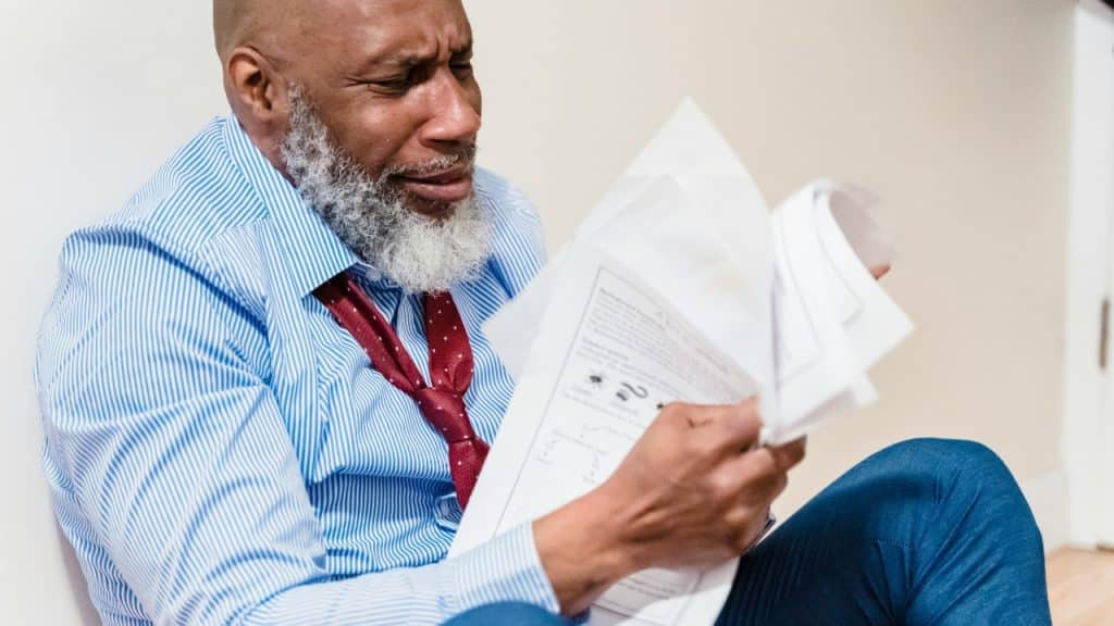A man sits on the floor, looking distressed while holding and reading several papers.