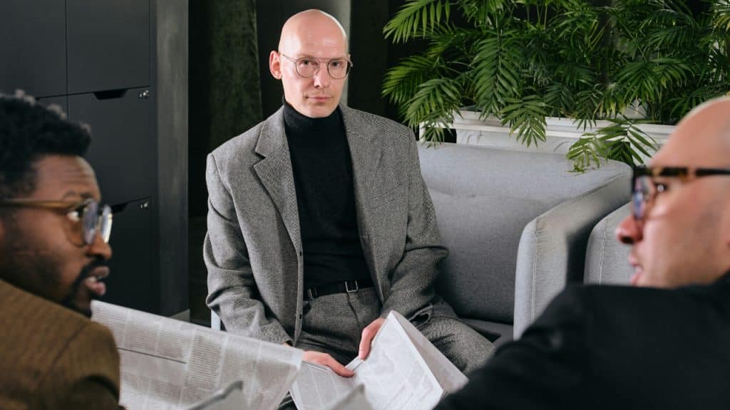A bald man in a gray suit sits on a sofa between two other men.