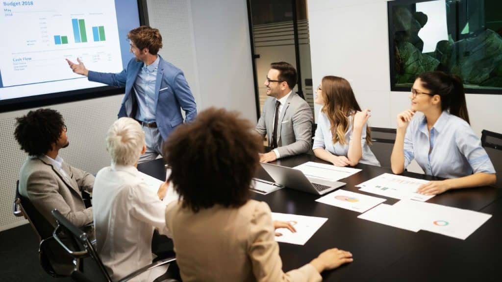 A man presents a budget chart to a group of colleagues around a table.
