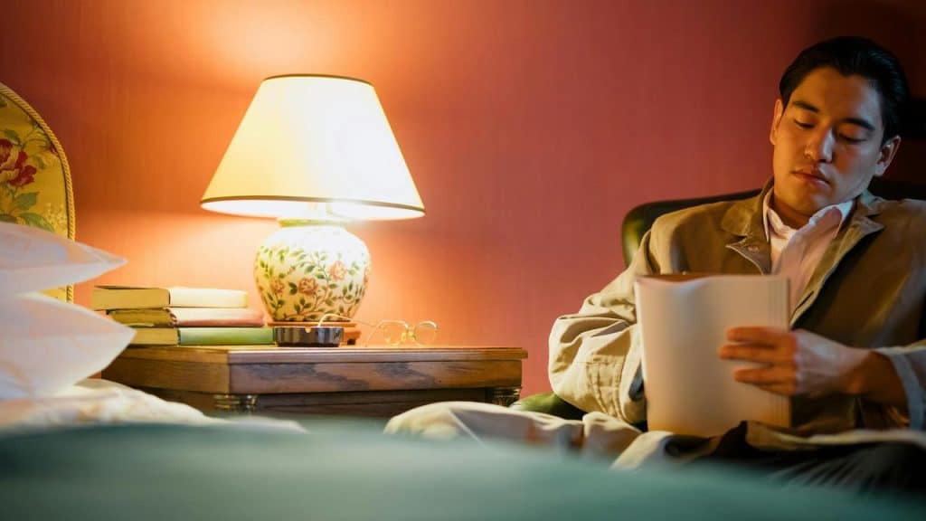 A man sitting in a chair reading papers beside a bedside table with a lamp and books.