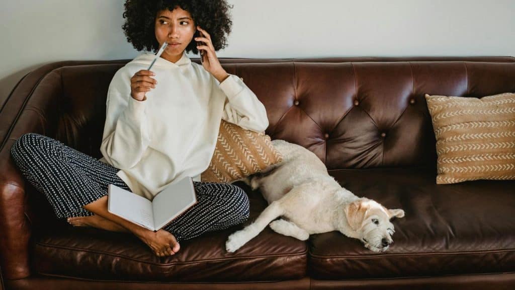 A woman sitting on a couch holding a pen with a dog beside her.