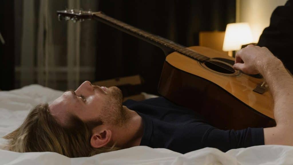A man lying on a bed holding an acoustic guitar in a dimly lit room.