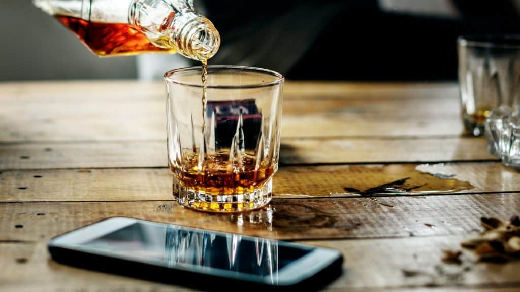 Amber liquid pours from a glass bottle into a small glass on a wooden table.