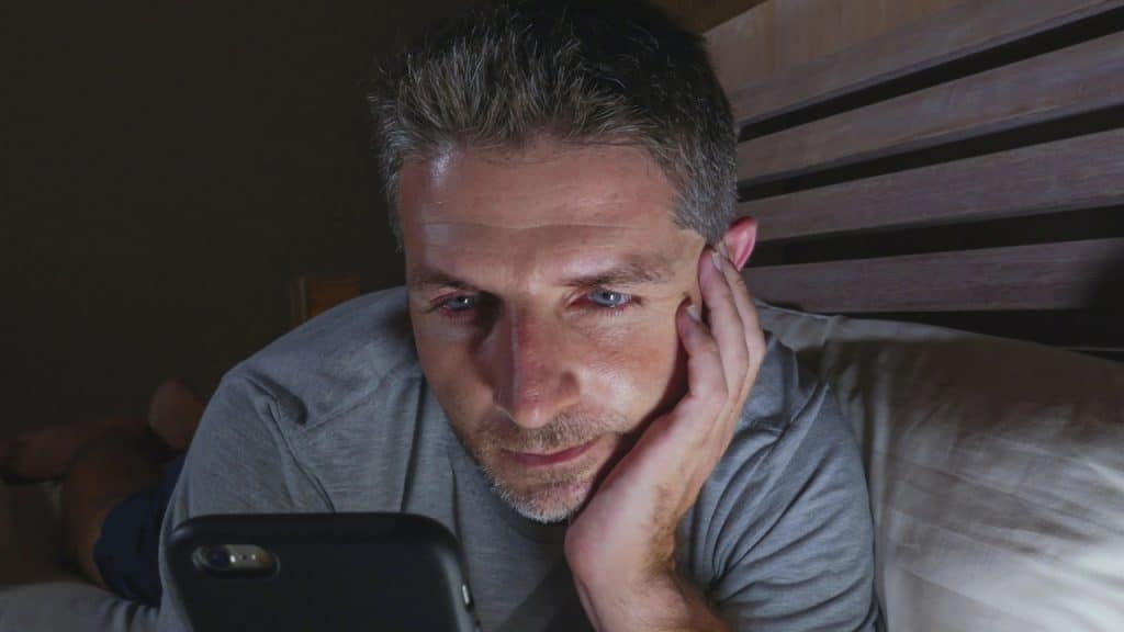 A man lies on a bed, looking down at his smartphone in the dark.