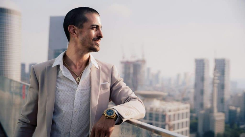A man in a light suit and gold necklace leans against a balcony overlooking buildings.