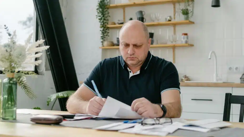 A bald man in a polo shirt sits at a table writing on paper.
