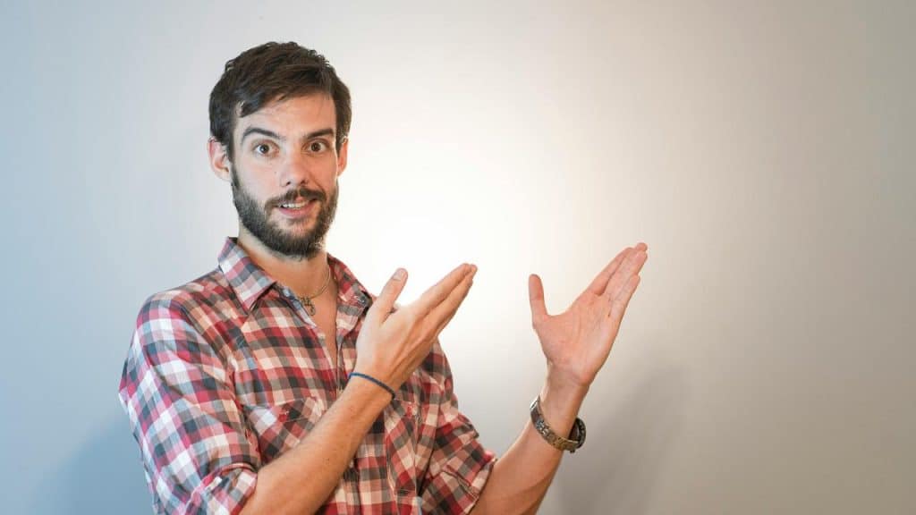 A surprised man in a plaid shirt gesturing with both hands toward empty space.