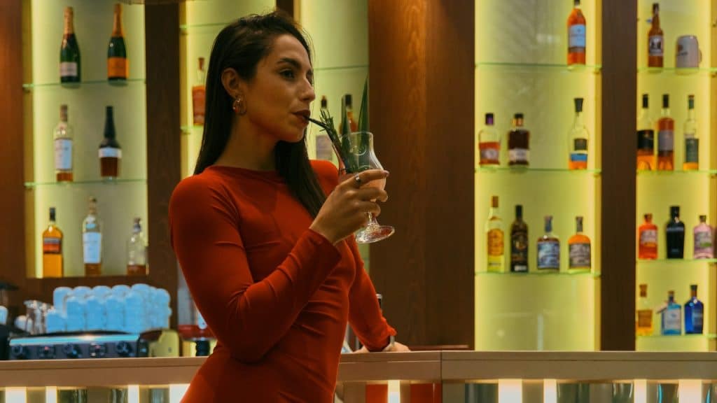 A woman in a red dress stands at a bar and drinks from a tall glass.