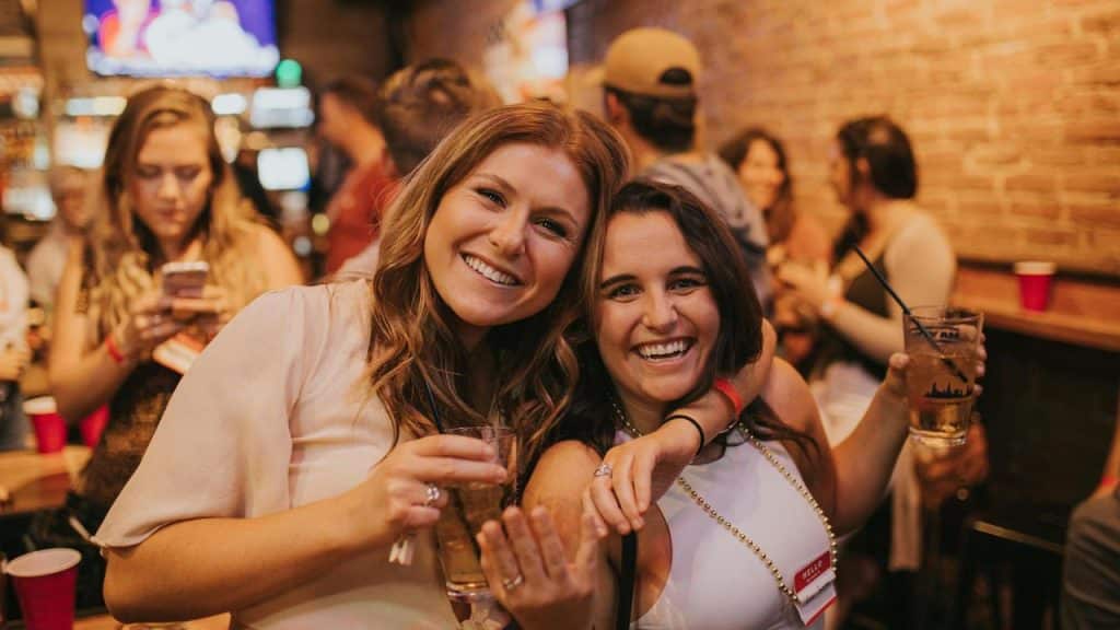 A smiling pair of friends holding drinks at a lively bar.