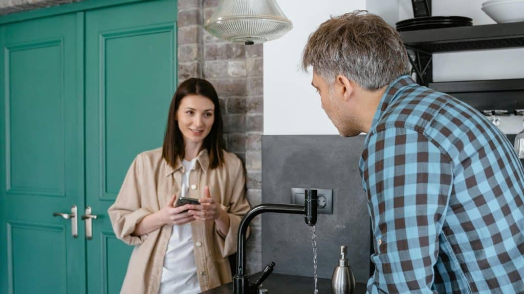 A woman holds a phone while looking at a man standing at a kitchen sink.