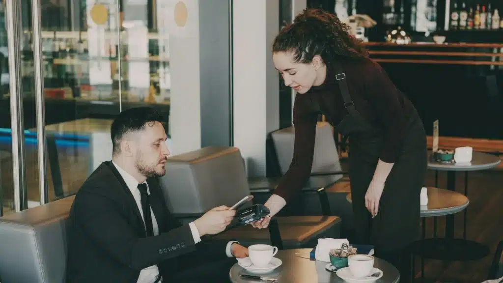 A man in a suit pays a server with his phone in a cafe setting.