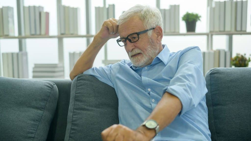 An older man with a white beard and glasses sits thoughtfully on a sofa.