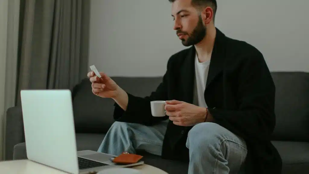 A man holding a cup and card while sitting with a laptop.