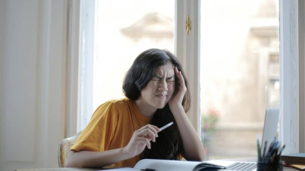 A woman holding her head in pain while sitting at a desk with a laptop.